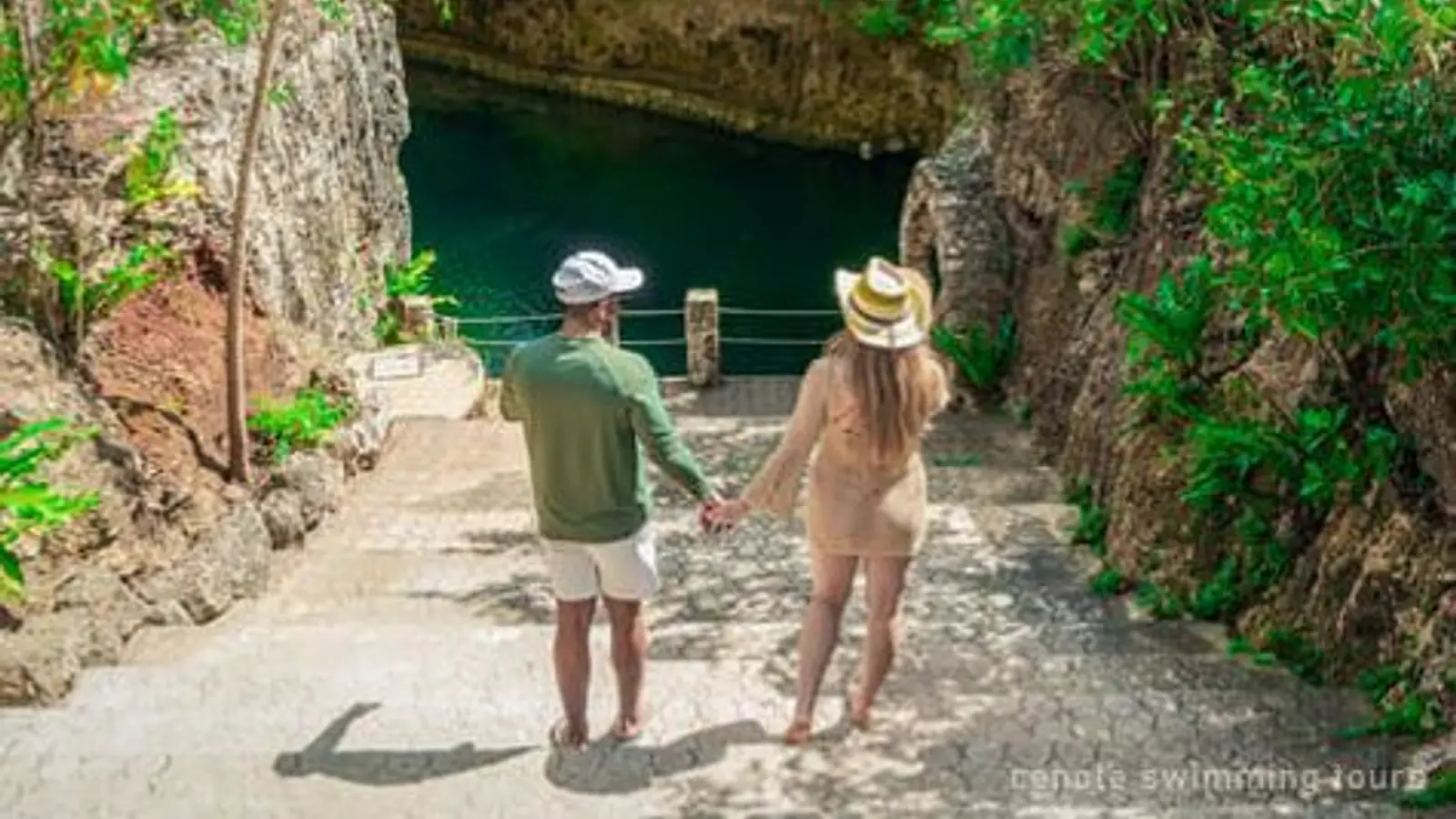 Two visitors walking down a path to a green cenote between rock walls — photo from the booking listing